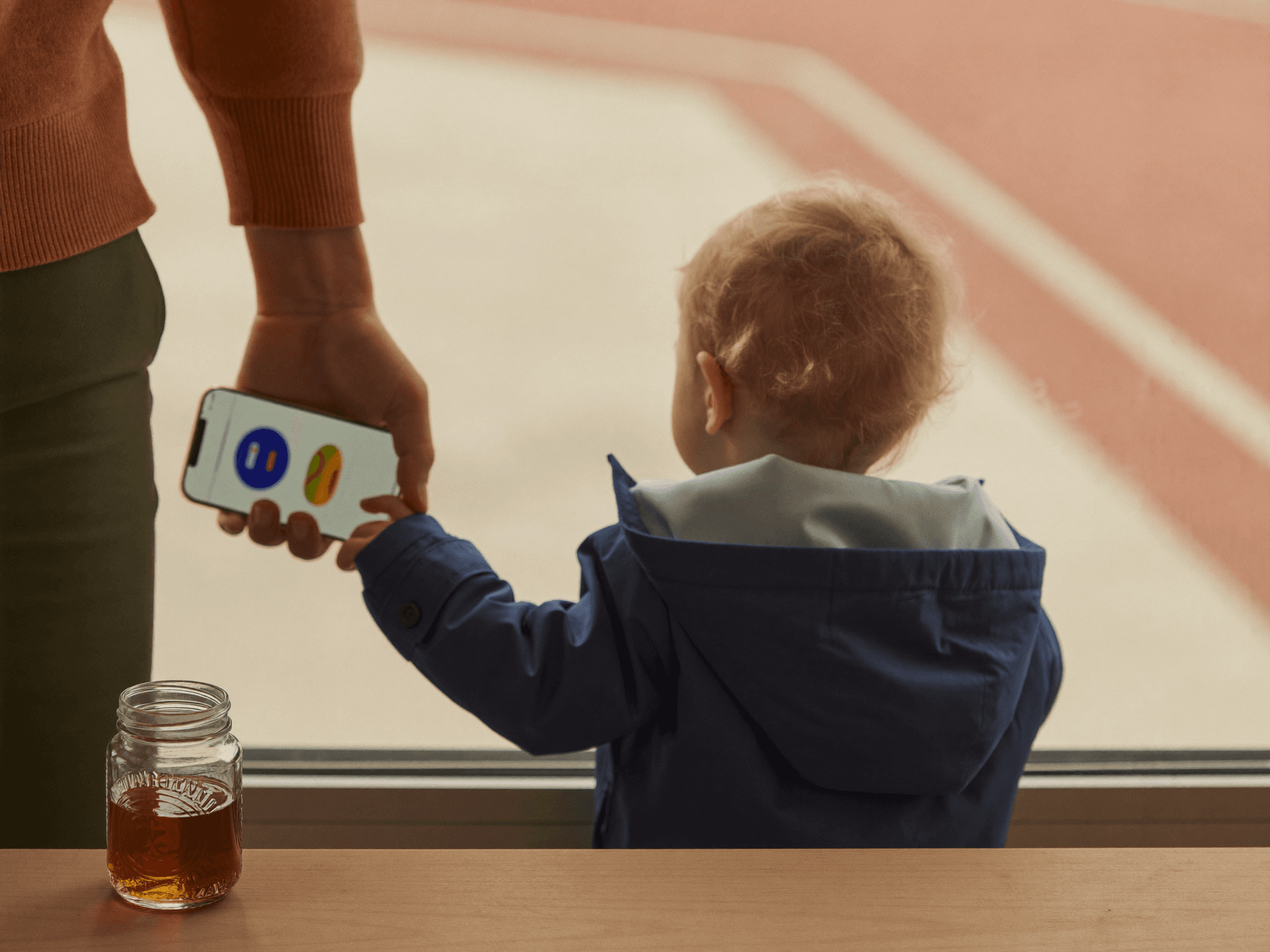 A parent holds a phone showing a customs lookup, a toddler in a navy raincoat stands beside them at an airport window, a jar of maple syrup on a wooden ledge.