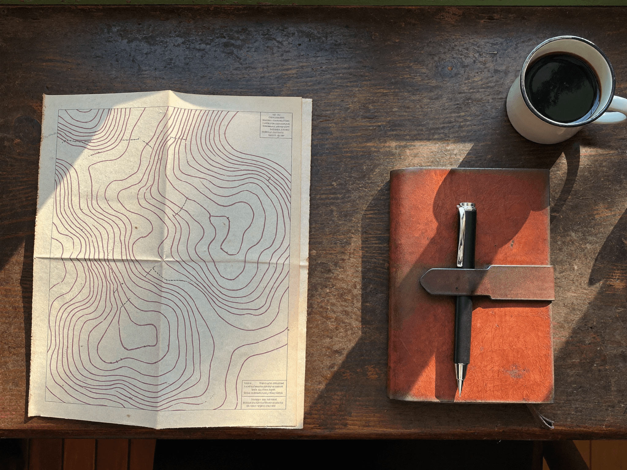 A vintage topographic map opened on a wooden desk beside a leather notebook with a fountain pen and an enamel mug of coffee.