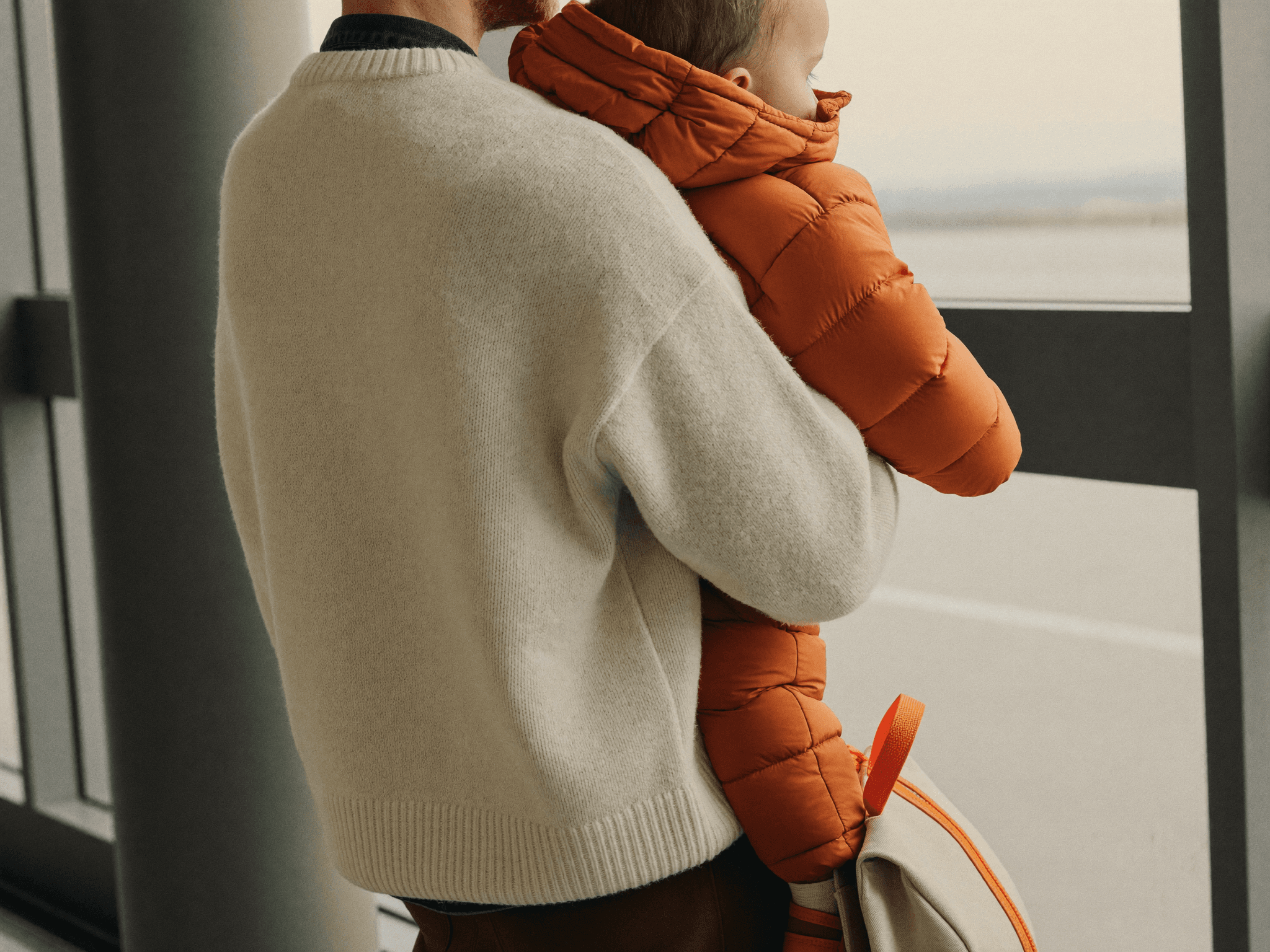 A parent in a cream wool sweater holds a toddler in a terracotta puffer jacket at an airport terminal window, viewed from behind.